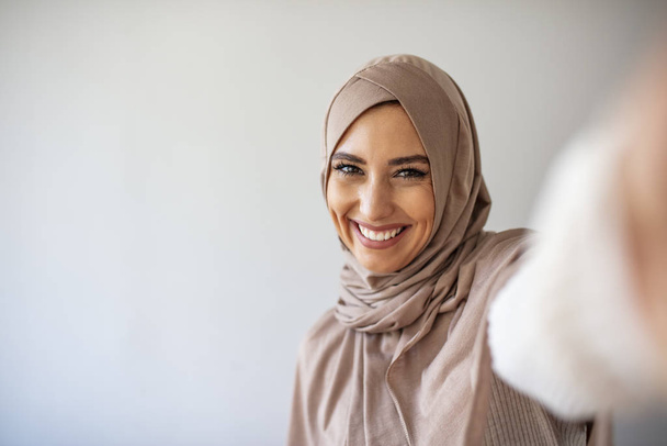 Arab beautiful woman smiling and selfie taking pictures by her mobile phone on gray backgound. Portrait of young muslim woman posing taking selfie photo with mobilephone - Foto, Imagem