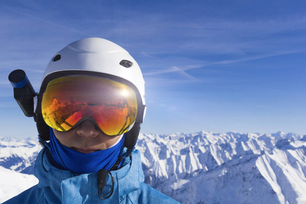 Skier άνθρωπος με πορτοκαλί γυαλιά σκι σε λευκό κράνος κάνουν φωτογράφηση στην κορυφή των Άλπεων βουνά. Στο φόντο των βουνών. Κλείσιμο προβολής. - Φωτογραφία, εικόνα