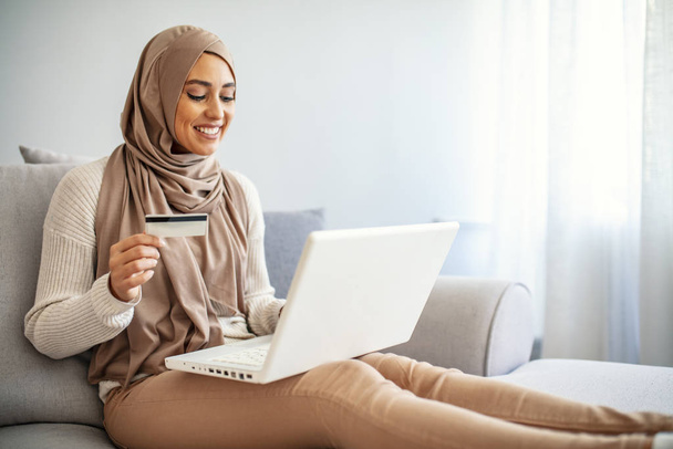 Die junge arabische Unternehmerin trägt einen Hidschab, arbeitet an einem Laptop und trinkt Kaffee, während sie im Wohnzimmer sitzt. Muslimin mit Laptop zu Hause. - Foto, Bild