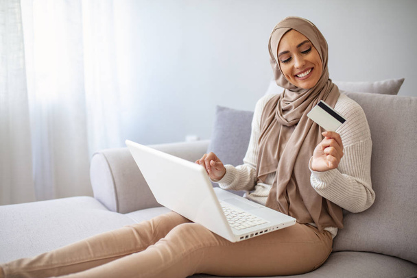 Portrait of beautiful young muslim woman browsing internet on laptop while holding a credit card at living room. Gorgeous young woman using laptop shopping online. - 写真・画像