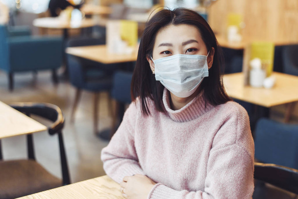 Asiatisches Mädchen mit Schutzmaske sitzt in einer Schule oder einem Café. Eine schöne Frau blickt in eine medizinische Maske in die Kamera, um nicht an das Virus zu erkranken. Prävention von Krankheiten - Foto, Bild