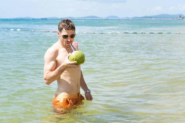 Summer vacation of handsome well shaped young man in sunglasses chilling on the blue sea and drinking coconut .Beautiful beach view with traveler man relaxing on Phu Quoc island, Vietnam.  - Foto, Bild