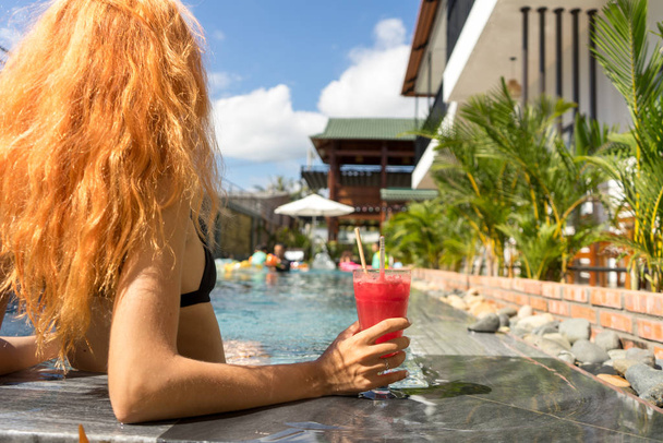 Estilo de vacaciones de verano pelirroja hermosa mujer sosteniendo jugo de sandía cerca de la piscina en vacaciones. Concepto de viaje
. - Foto, imagen