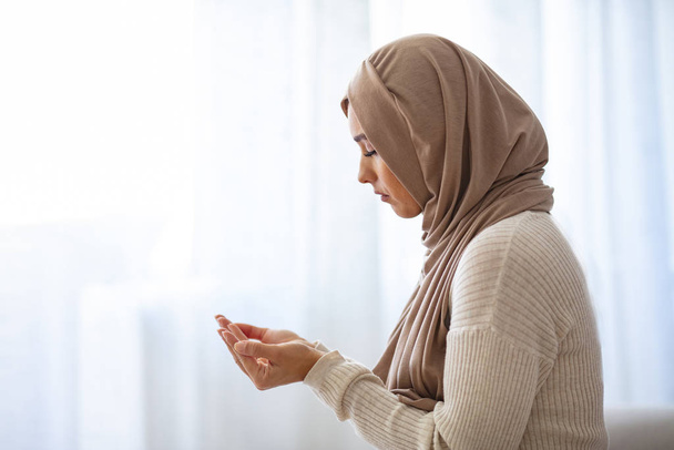 Muslim woman in beige hijab and traditional clothes praying for Allah, copy space. Muslim woman with hijab praying indoor at bright window. Young Muslim woman Praying - Fotoğraf, Görsel