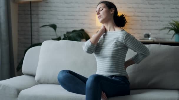 Video of young woman with back and neck pain sitting on the sofa in the living room at home. - Materiał filmowy, wideo
