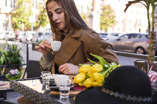 Jong mooi meisje drinkt espresso in een café op straat. Voorjaarszon in beeld. Er ligt een boeket bloemen op tafel. Ochtendpauze voor een kop koffie. Kopieerruimte. - Foto, afbeelding