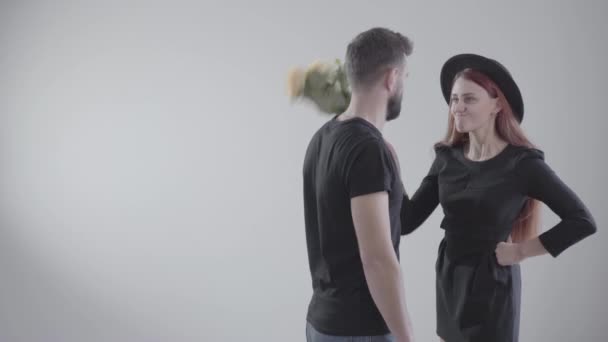 Elegant offended Caucasian girl in black dress and hat beating her boyfriend or husband with flowers. Young handsome man leaving the shot with bouquet. Conflict of young couple at white background. - Footage, Video