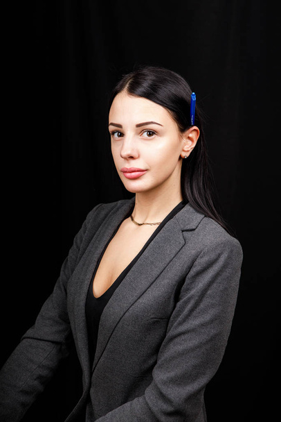 Portrait of young business woman in  a grey jacket on black background with pen behind the ear - Фото, изображение