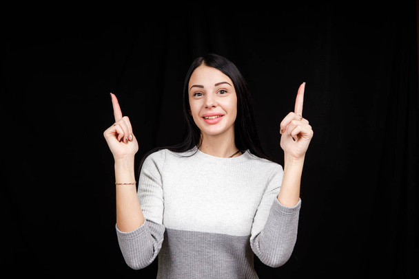 The concept of advertising. Positive, pretty, caucasion woman with a beaming smile and bright sweater on a black background indicates two index fingers and looks at the empty copy space  - Fotoğraf, Görsel