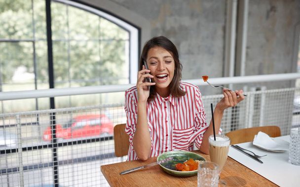 beautiful young brunette talking on a cell phone in restaurant while eating. - Photo, image