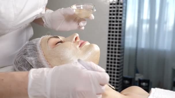 Beauty concept. Beautician Applying cosmetic golden mask on beautiful female face skin. Face masks usage in modern cosmetology. Portrait of young girl getting facial procedure in beauty salon. 4 k - Footage, Video