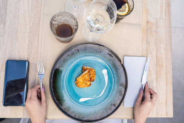 Luchtfoto van een blauwe schotel met stukjes gebakken kip met yoghurtsaus als decoratie van een minimalistisch schaaltje met glazen water en frisdrank en een mobiele telefoon op tafel terwijl er handen met mes en vork klaar zijn om te eten - Foto, afbeelding