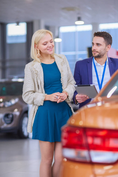 Dealer with woman stands near a new car in the showroom - Photo, image