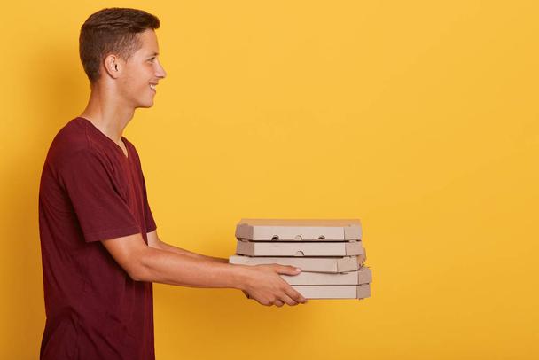 Side view of delivery man wearing maroon casual t shirt giving food order pizza boxes isolated over yellow studio background. Service and delivery concept. Copy space for advertisment or promotion. - Fotó, kép