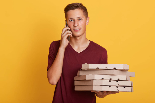 Indoor shot of attractive male wearing butgundy casual t shirt, guy talking to phone with his client, holding pizza boxes in hands, standing isolated over yellow studio background. Delivery concept. - 写真・画像