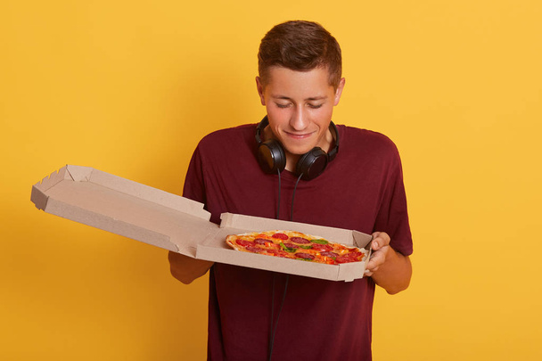 Close up portrait of handsome guy wearing burgundy t shirt smelling fresh pizza from carton box and gets pleasure, standing isolated over yellow background, young man being ready to eat junk food. - Fotoğraf, Görsel