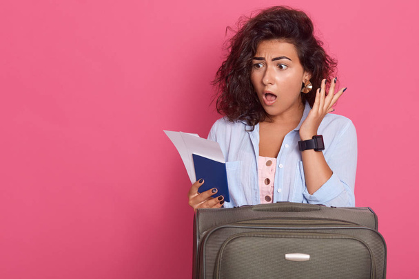 Close up portrait of young brunette woman posing isolated on pink background, being ready to have vacation, stands with suitcase, tickets and passport, looks surprised, poses with widely opened mouth. - Fotoğraf, Görsel
