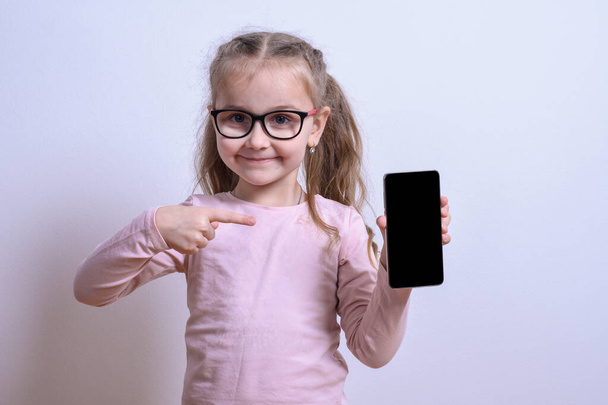 Egy fehér kislány portréja szürke alapon mutat a telefonra. - Fotó, kép