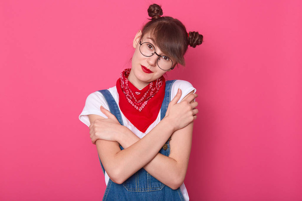 Close up portrait of attractive woman wearingdenim overalls, white casual t shirt, red bandana over neck, posing isolated over pink background, female looking directly at camera, hugging herself. - Фото, изображение