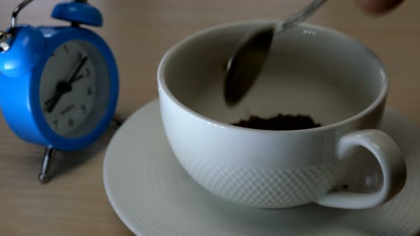 Making morning coffee. Instant coffee spread on in white cup with a teaspoon, poured with boiling water and mixed. Blue alarm clock is standing and ticking near. Start of day concept. - Πλάνα, βίντεο