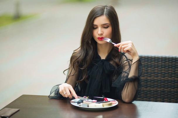 beautiful young woman in a black dress with makeup sits at a table with cosmetics in a plate - Фото, изображение