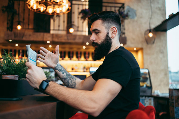 Young adult hipster man with a long beard sitting in modern cafe bar, reading newspapers and emotionally reacting. - Foto, Imagem