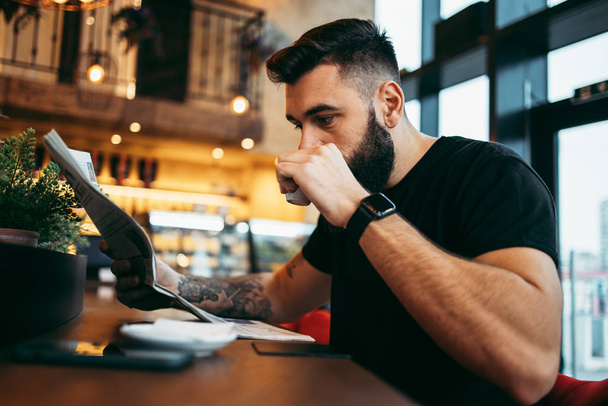 Young adult hipster man with a long beard sitting in modern cafe bar, reading newspapers and emotionally reacting. - 写真・画像