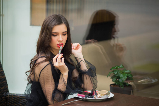 beautiful young woman in a black dress with makeup sits at a table with shadows, lipstick, mascara in a plate - Photo, image
