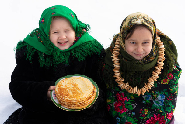 Two charming girls in a Russian folk costume sit on the snow and eat pancakes. The concept of the celebration of the Russian holiday shrovetide. - Photo, Image