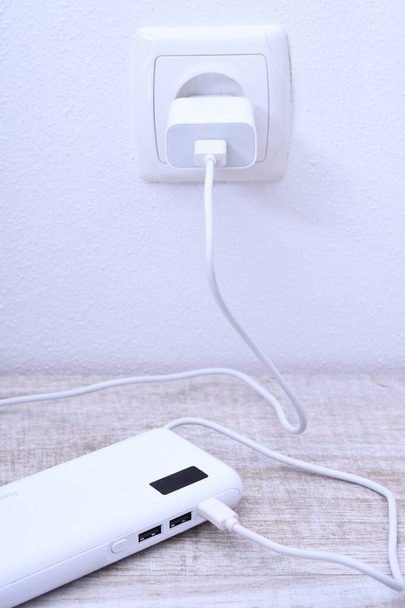 Biały telefon power bank podłączony do gniazdka na ścianie do ładowania na drewnianym tle - Zdjęcie, obraz