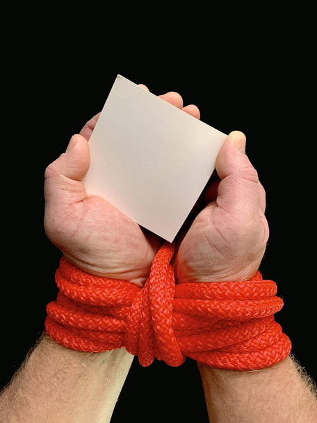 Manos atadas con una cuerda, sobre un fondo negro. Las manos atadas sostienen una nota con la inscripción. Concepto: préstamo, deuda, ayuda
. - Foto, imagen