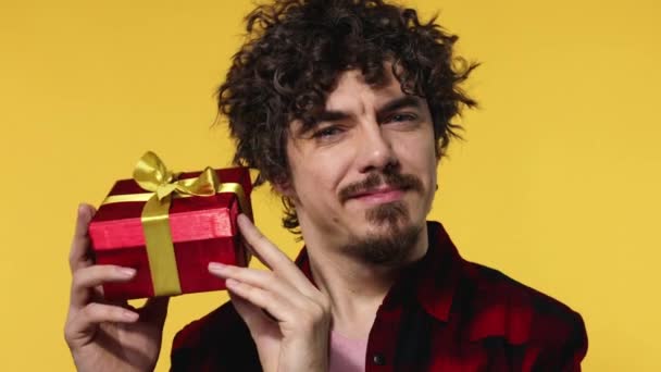 A man gives a red gift for Valentines Day or International Womens Day. Birthday surprise. Handsome happy european man with beard in shirt smiling isolated on yellow background. - Imágenes, Vídeo