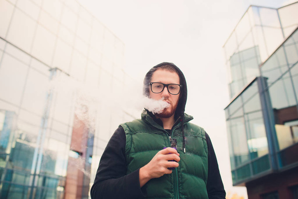 Portrait of young guy with large beard in glasses vaping an electronic cigarette opposite urban background. - Фото, изображение