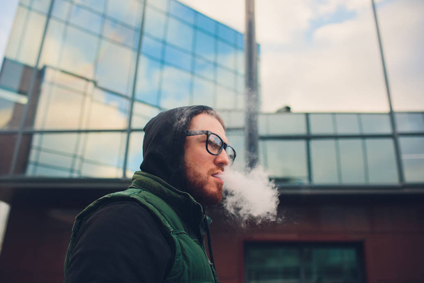 Portrait of young guy with large beard in glasses vaping an electronic cigarette opposite urban background. - Photo, image