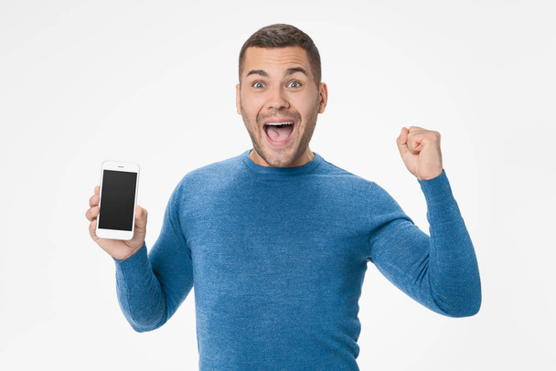 Portrait of happy young man holding mobile phone and celebrating success isolated over white background - Photo, Image
