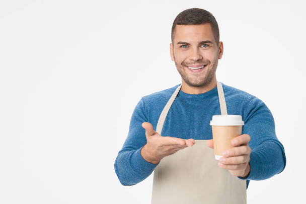 Positive handsome man holding paper cup recommending you to taste it wearkng apron isolated over white background - 写真・画像