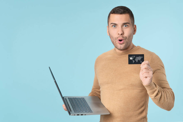 Portrait of surprised handsome man wearing casual clothes using laptop computer and showing credit card standing isolated over blue background - 写真・画像