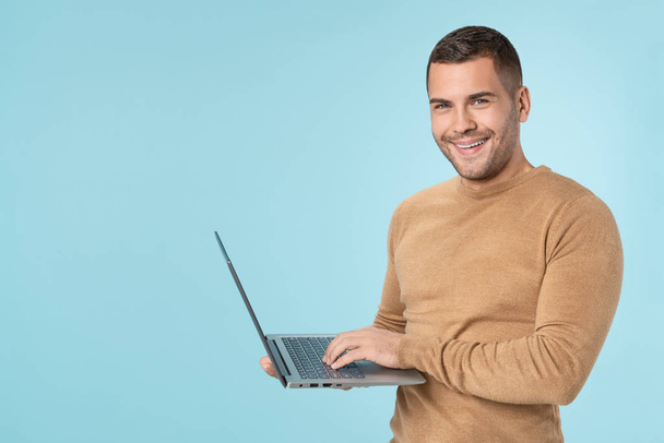 Happy young man using laptop and looking at camera over blue background - Zdjęcie, obraz