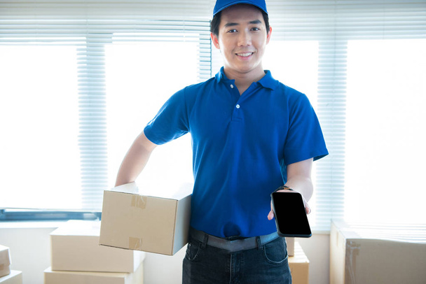 Young delivery man in uniform holding and showing smartphone with blank screen.Ai transportation technology concept. - Photo, Image