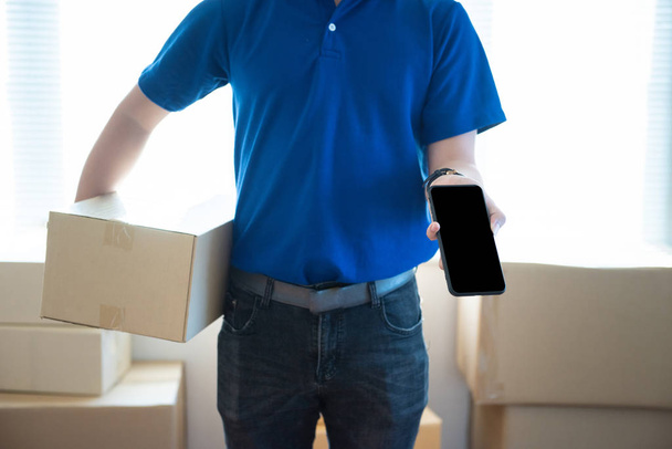 Young delivery man in uniform holding and showing smartphone with blank screen.Ai transportation technology concept. - Photo, Image