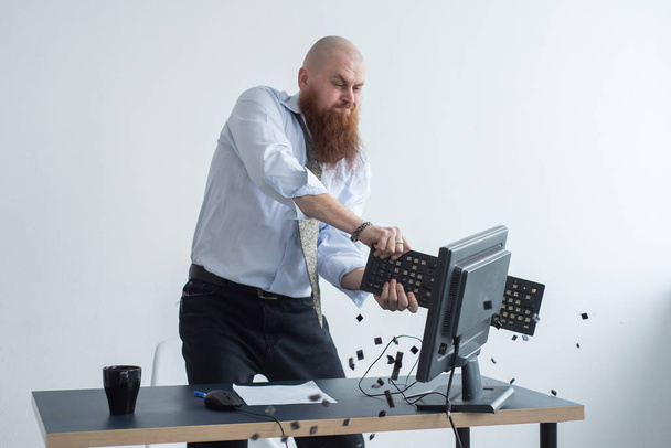 Angry bald man with a red beard in the office in a business suit crashes a computer. Manager with a nervous breakdown breaks the keyboard on the monitor. - Foto, Imagem