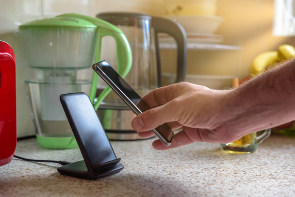 ασύρματη φόρτιση smartphone με βάση φόρτισης στο τραπέζι της κουζίνας. αρσενικό χέρι τοποθέτηση του τηλεφώνου στη φόρτιση - Φωτογραφία, εικόνα