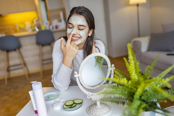 So beautiful. Close-up of girl with beauty mask on her face looking in mirror. Girl uses natural mask. Attractive young woman sitting with a facial mask on her skin - Valokuva, kuva