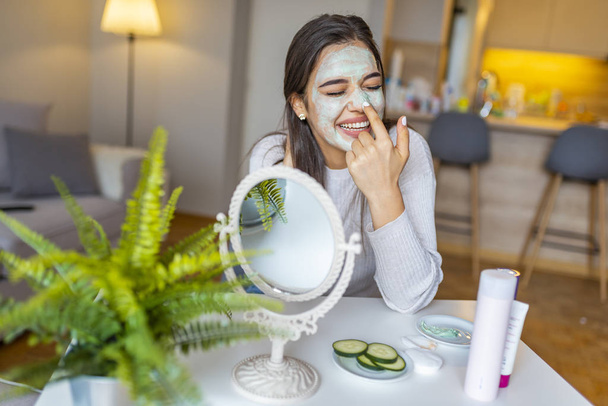 So beautiful. Close-up of girl with beauty mask on her face looking in mirror. Girl uses natural mask. Attractive young woman sitting with a facial mask on her skin - Photo, image