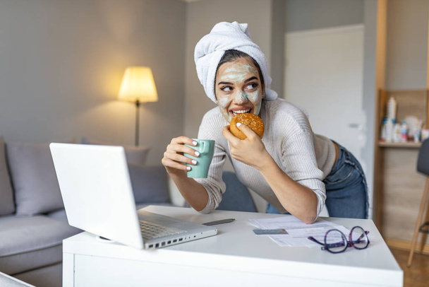 Beautiful young woman working on laptop computer while sitting at the living room, drinking coffee with face mask. Beautiful young woman smiling with face mask and looking at laptop screen - Foto, Imagen