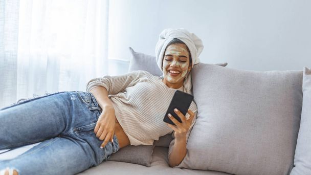 Young woman with white clay facial mask using smartphone while sitting on bed at home. Woman having face treatment at home lying on grey bed. Woman use beauty mask with cellphone at home - Photo, Image