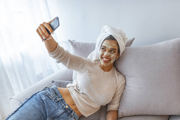 Close up selfie of beautiful happy woman after shower with a towel on her head, with cream on face. Woman with clay mask taking selfie with mobile phone at home enjoying relaxation  - Foto, afbeelding