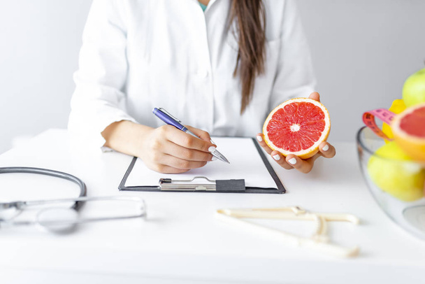Nutritionist desk with healthy fruit, juice and measuring tape. Dietitian working on diet plan. Weight loss and right nutrition concept. Unrecognizable woman dietitian typing, counting calories or writing diet plan, copy space. - Fotó, kép