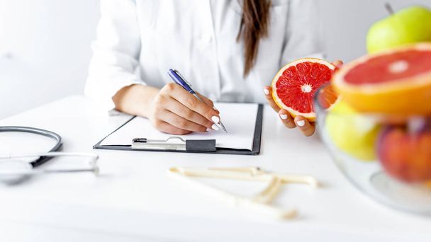 Doctor nutritionist writing case history in the office. Young woman dietitian prescribing recipe. Female nutritionist sitting at table with clipboard and healthy products on white background - Photo, image