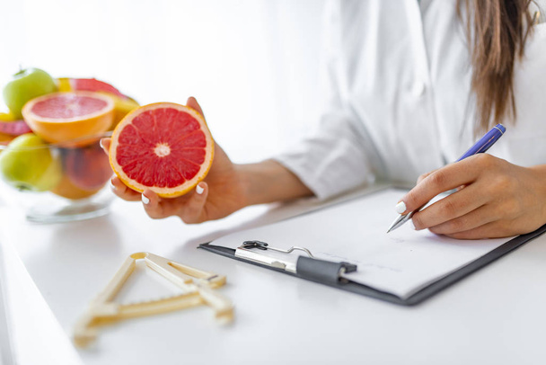 Doctor nutritionist writing case history in the office. Young woman dietitian prescribing recipe. Female nutritionist sitting at table with clipboard and healthy products on white background - Φωτογραφία, εικόνα
